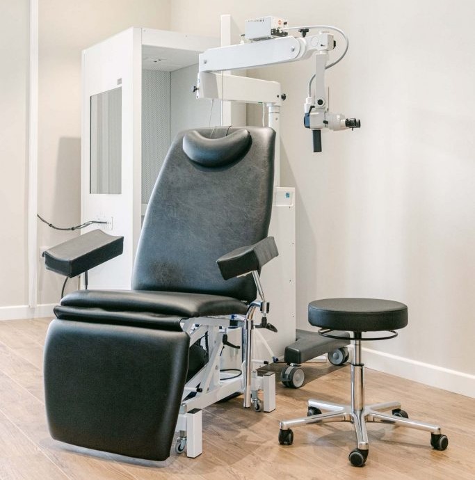 A dark padded examination chair and wheeled stool sit beneath an adjustable examination scope in a bright clinic room.