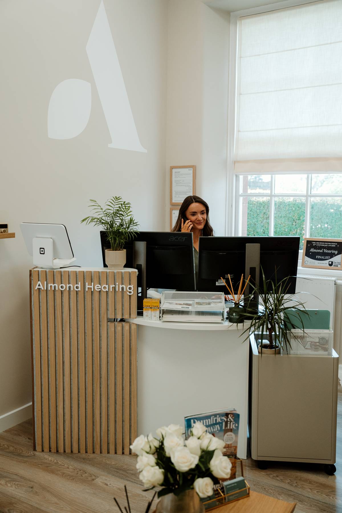 A receptionist speaks on the phone behind the Almond Hearing desk, surrounded by computer screens, potted plants and a sunlit window.