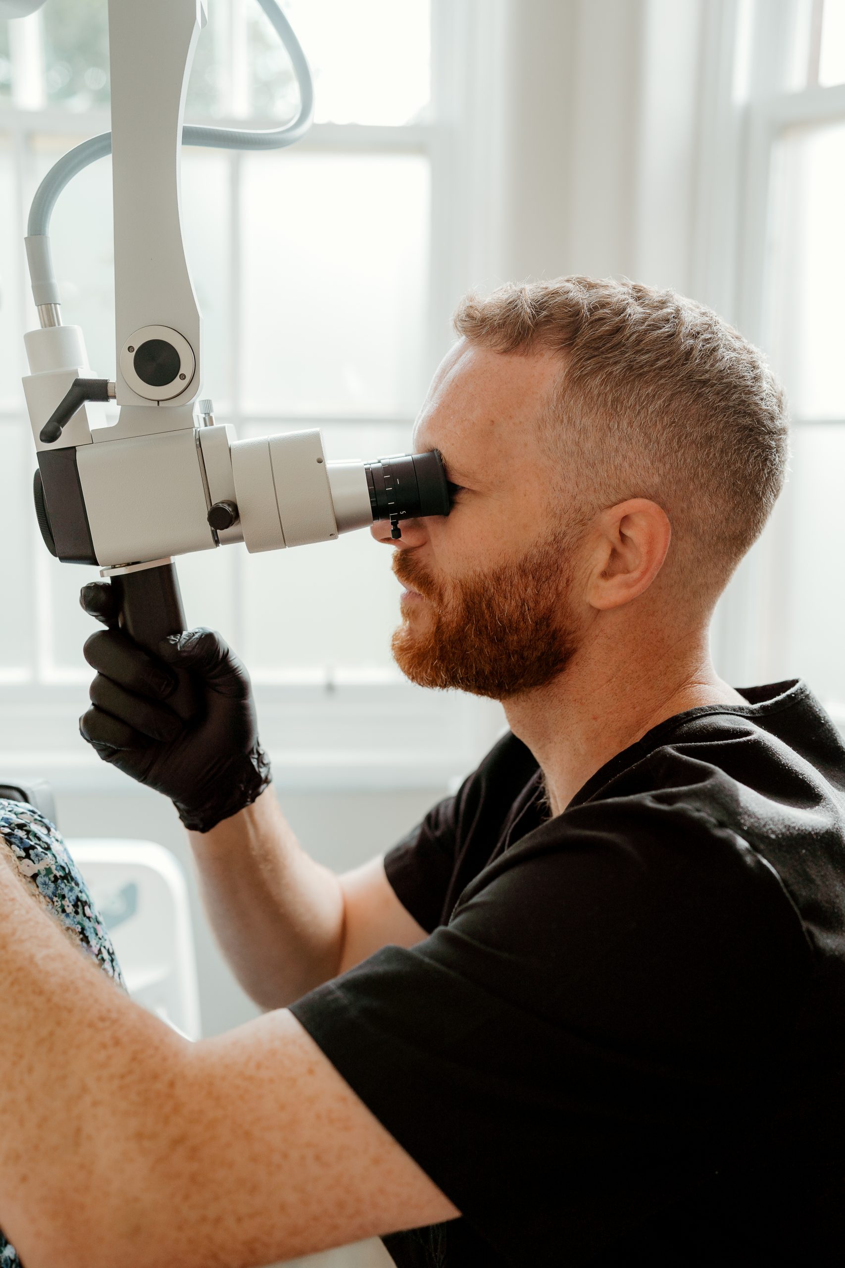A clinician in black gloves examines a patient through a microscope during a detailed hearing assessment at an Almond Hearing clinic.