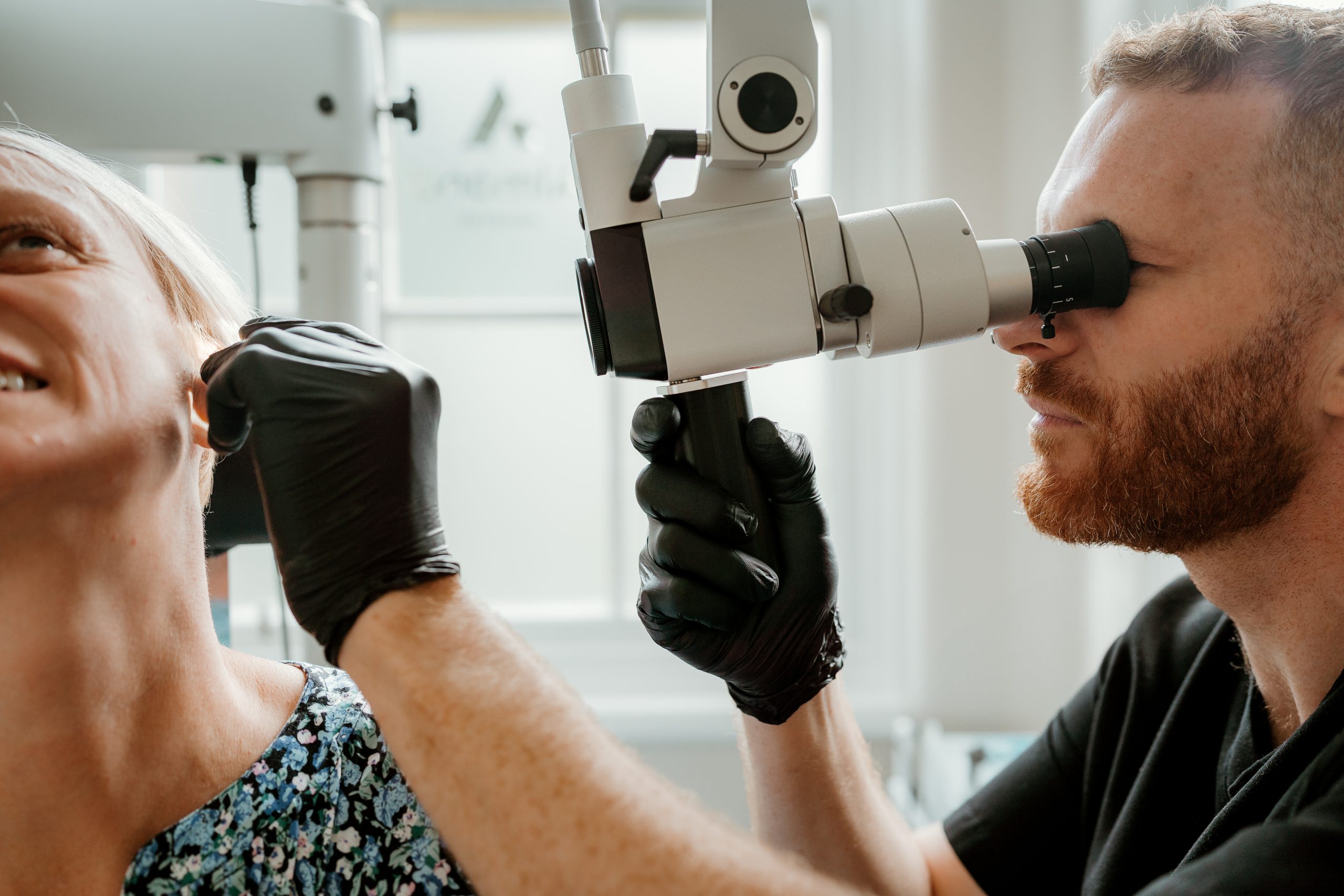 A clinician in black gloves examines a patient’s ear through a high-powered microscope during a hearing clinic appointment.