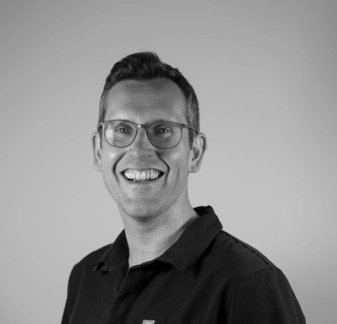 A smiling man with short dark hair and glasses wears a black collared shirt in a monochrome portrait of vestibular audiologist Peter Lucas-Herald.