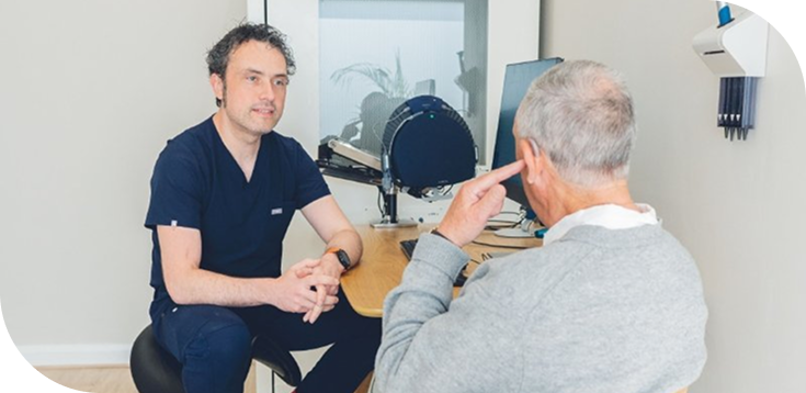 An audiologist in navy scrubs talks with an older patient in a grey jumper beside hearing assessment equipment in a clinic.