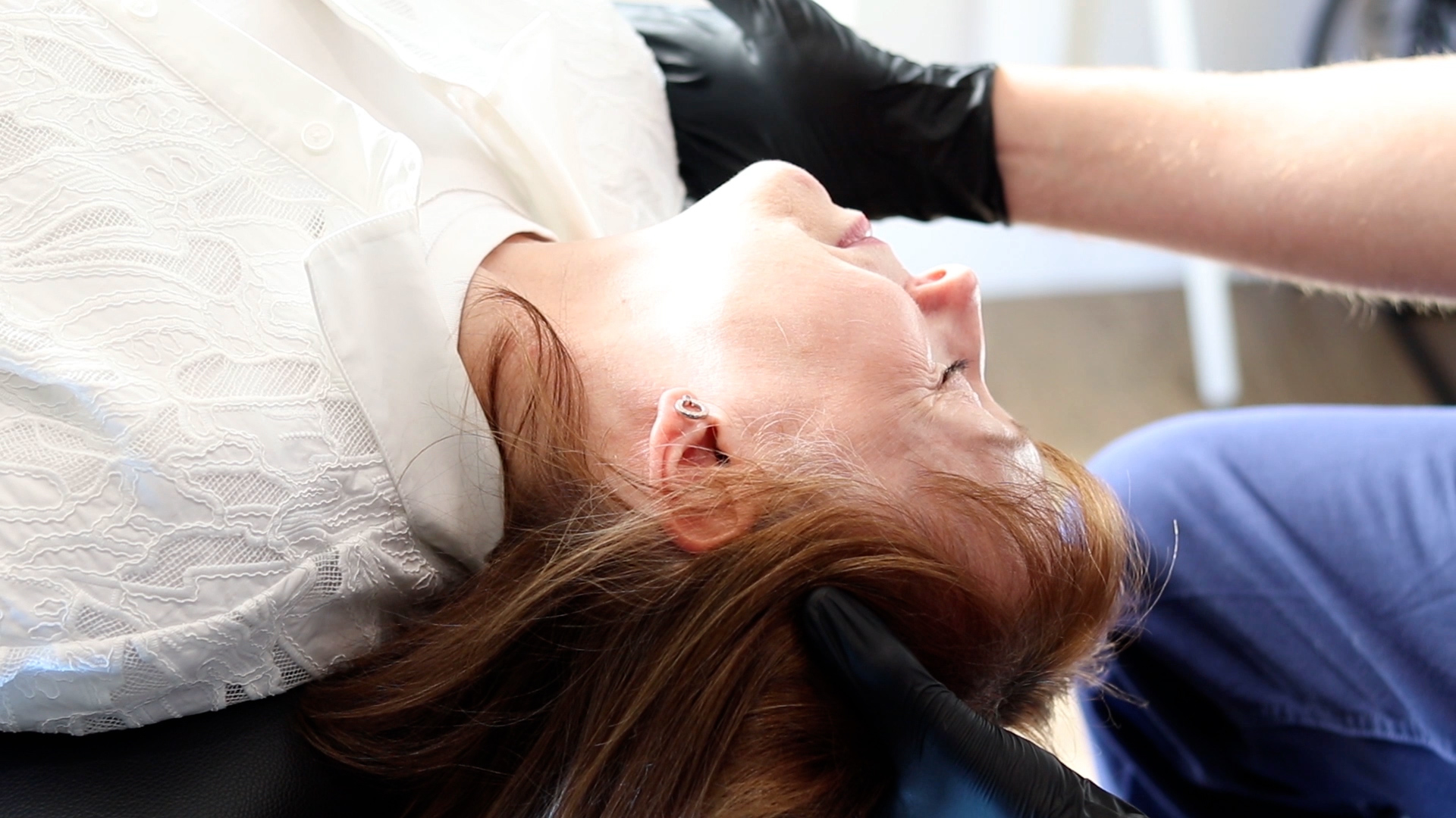 A woman reclines with eyes closed while a gloved clinician gently supports her head during a balance assessment at a hearing clinic.