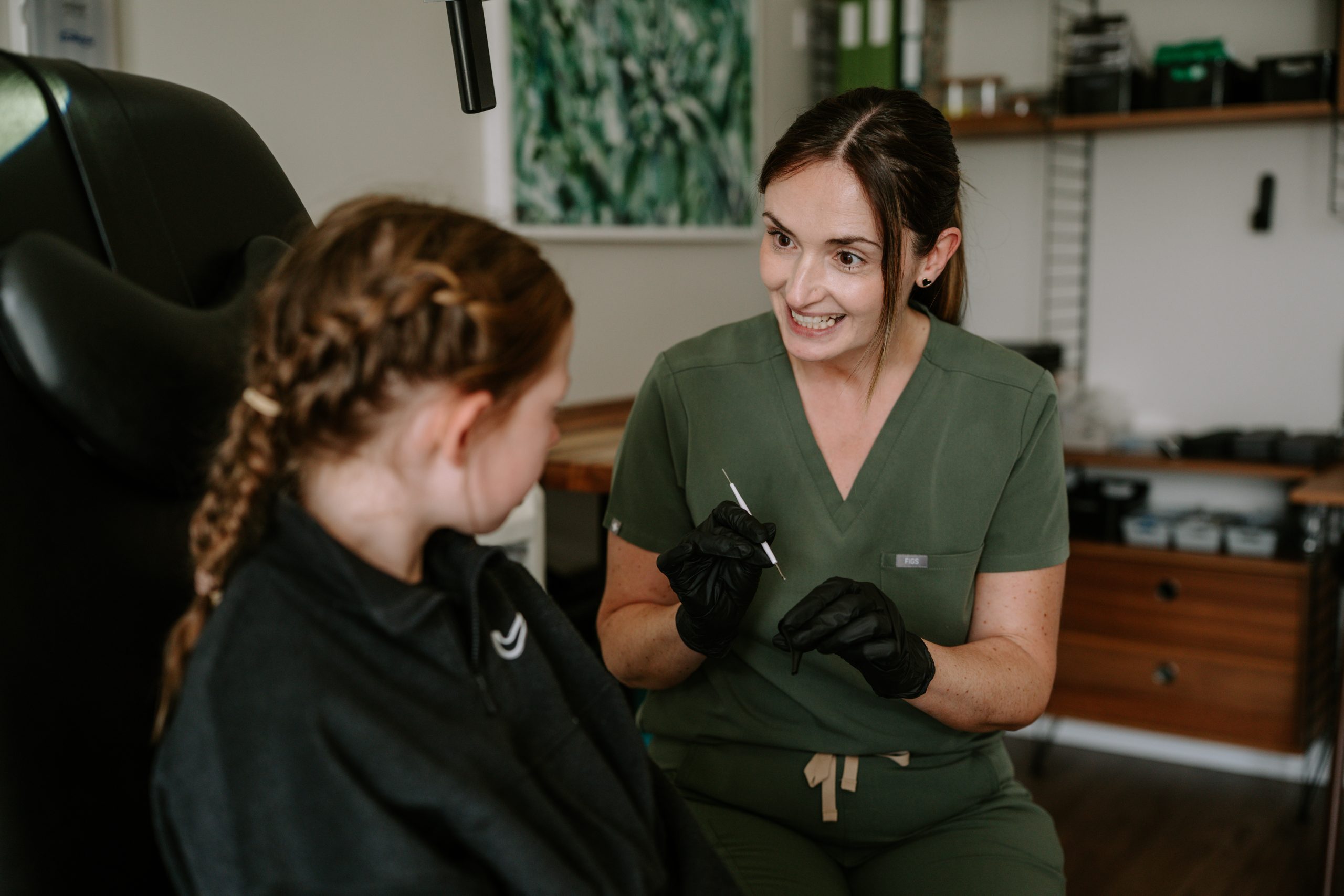 In an Almond Hearing clinic in Scotland, a clinician in green scrubs and black gloves speaks with a seated young patient.