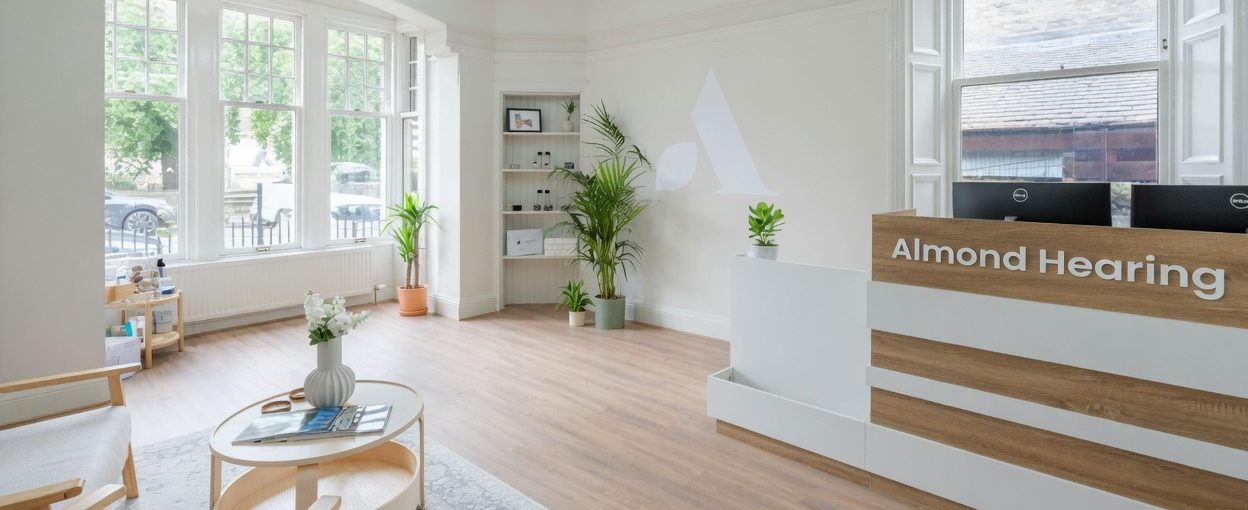 Bright Almond Hearing reception in Broughty Ferry, with a wood-and-white desk, tall windows, pale flooring and green plants.