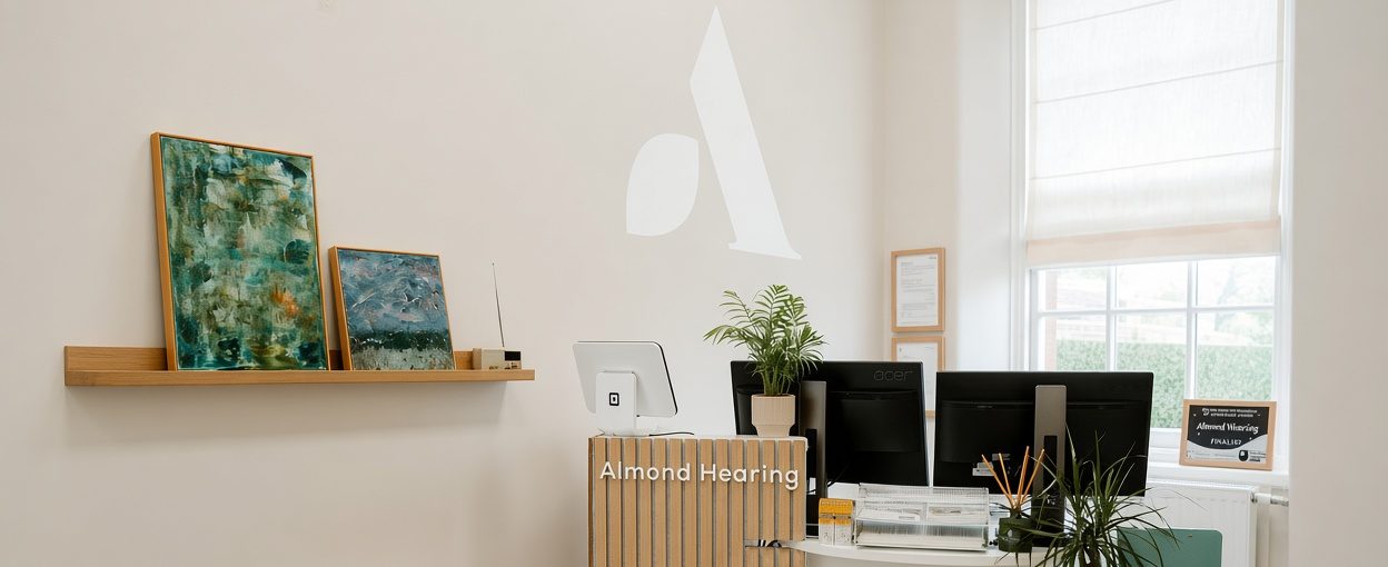 The Almond Hearing reception in Dumfries has a timber-slat desk, framed artwork, plant and computer monitors beside a bright window.