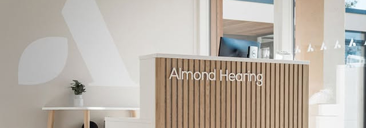 A light wood reception desk with the Almond Hearing name stands in a bright, modern clinic interior in Livingston, Scotland.