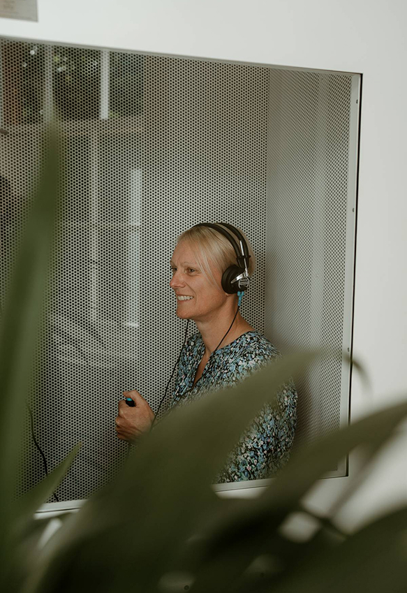 A smiling blonde woman wears headphones and holds a control in a soundproof booth during a hearing assessment at Livingston Hearing Clinic.