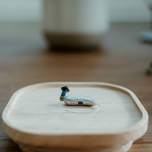 A silver behind-the-ear hearing aid rests on a pale wooden tray, with its ear hook visible against a softly blurred background.