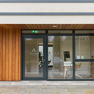 The Almond Hearing clinic in Livingston has a glass-fronted entrance with dark frames, warm timber cladding and a view inside.