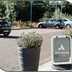 A grey Almond Hearing sign and leafy potted plant stand outside the Livingston clinic, with parked cars and trees behind them.