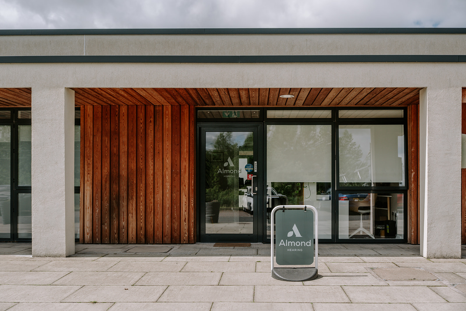 Almond Hearing’s Livingston clinic has a glass-fronted entrance framed by warm timber cladding, with a branded pavement sign outside.