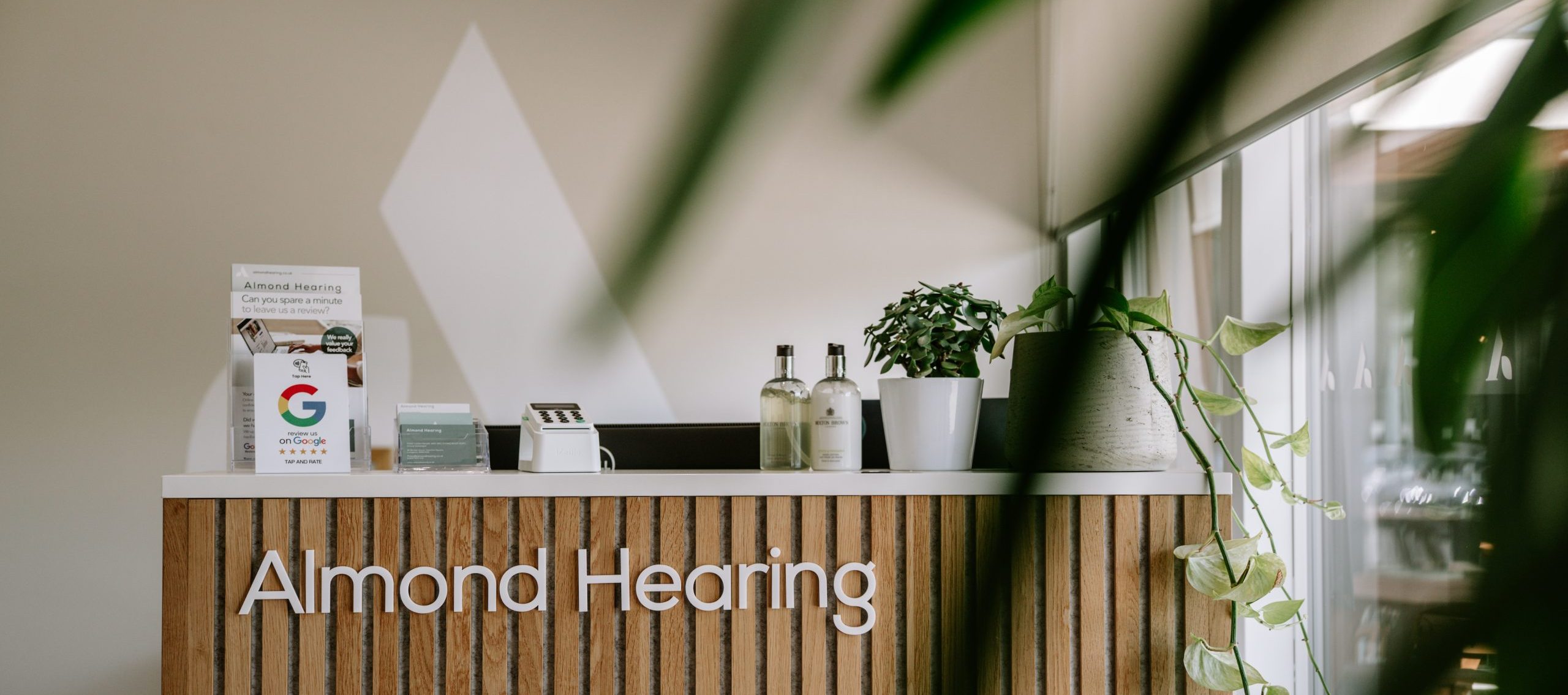 Almond Hearing’s Livingston reception has a slatted wooden desk, review cards and potted plants beside a bright window.