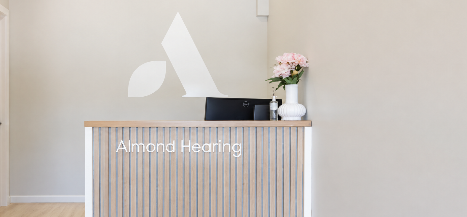 Wooden slatted reception desk with the Almond Hearing name, a black monitor, hand sanitiser and pink flowers in a white vase.