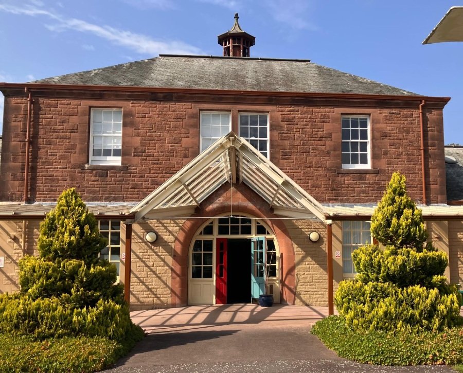 A Dumfries clinic has a red sandstone façade, a steep glass entrance canopy, open red and blue doors, and clipped shrubs.