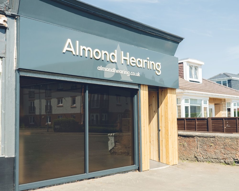 The Clarkston Almond Hearing clinic has a teal sign above dark glazed doors, pale timber cladding and a sunlit pavement.