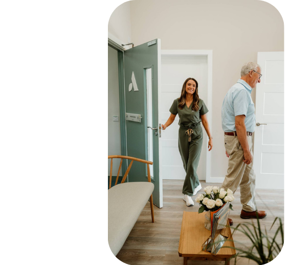 A smiling audiologist in green scrubs welcomes an older man into a bright clinic room, with a chair and flowers nearby.