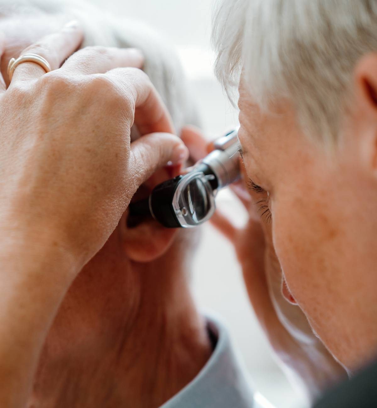 A clinician examines an older adult’s ear with a silver handheld otoscope, bringing the instrument close to the ear canal.