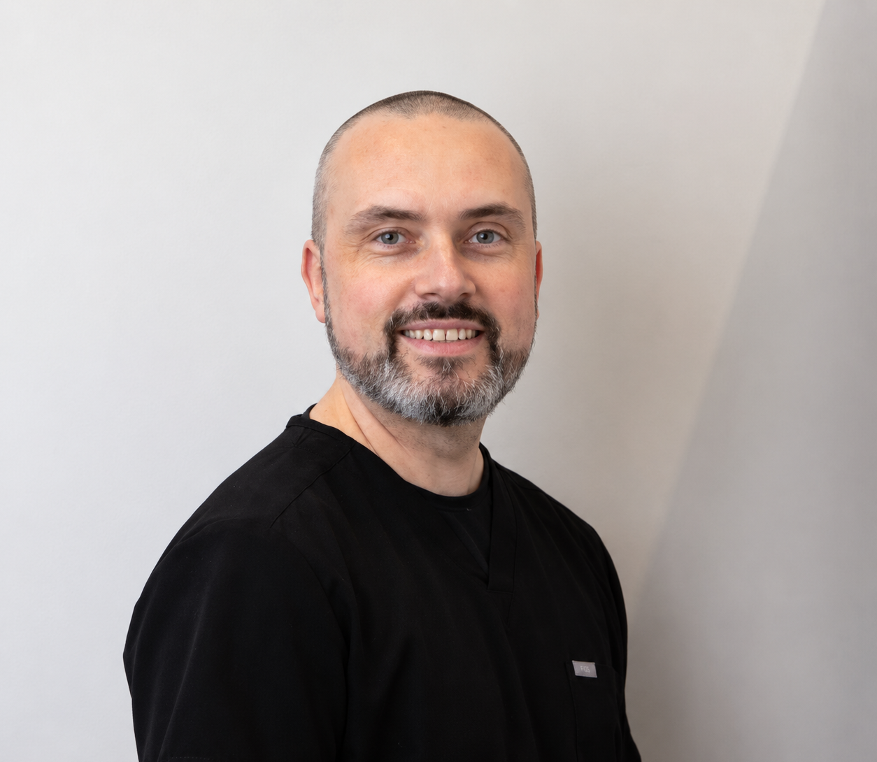 Smiling audiologist Iain has close-cropped hair and a salt-and-pepper beard, wearing black clinical clothing against a pale backdrop.